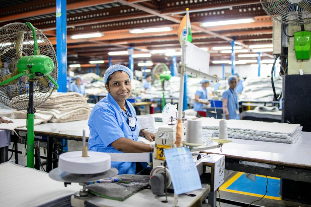 A female factory worker in India manages a sewing machine in a bustling textile factory.